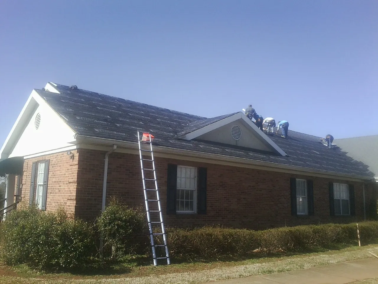 Crew tearing shingles off a brick ranch roof with a ladder set — Metal Roof Repair work by our crew in Archie