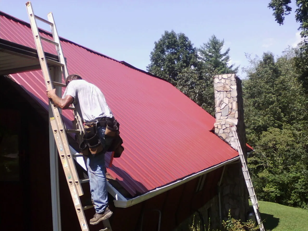 Roofer on a ladder beside a red metal roof and stone chimney — Soffit & Fascia Repair by Roofing Brothers in Archie